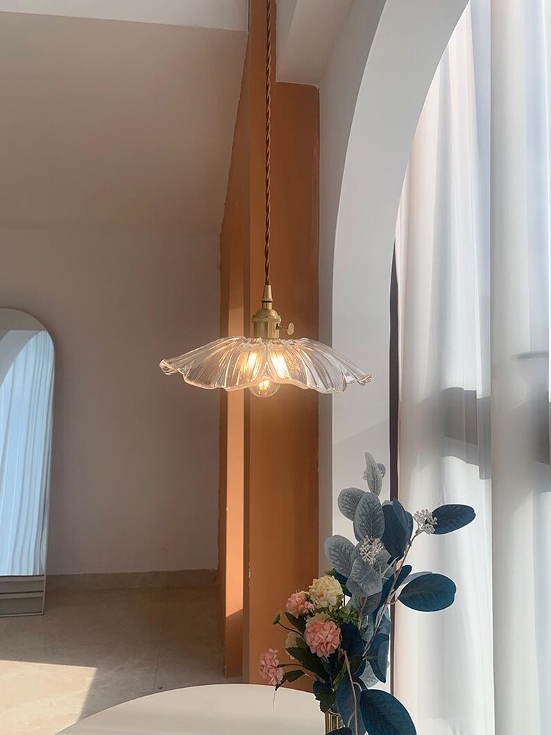 May include: A clear glass pendant light with a ruffled edge, hanging from a brown cord. The light fixture has a brass-colored base and is illuminated. The background includes a vase of flowers and a window with white curtains.