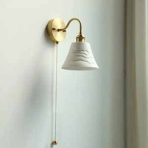 May include: A gold wall-mounted light fixture with a white ceramic shade featuring a textured pattern. The light has a pull chain for turning it on and off.