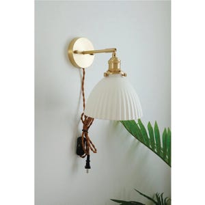 Op de afbeelding: Een witte keramische wandlamp met een gouden metalen arm en voet. De lamp heeft een geplooide kap en een bruin snoer met een zwarte schakelaar.