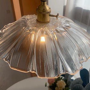 May include: A clear glass pendant light fixture with a ruffled, flower-like shade. The light fixture has a brass-colored base and a visible lightbulb. The shade has a textured surface and is suspended from the ceiling.