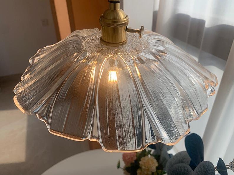 May include: A clear glass pendant light fixture with a ruffled, flower-like shade. The light fixture has a brass-colored base and a visible lightbulb. The shade has a textured surface and is suspended from the ceiling.