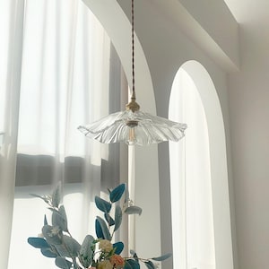 May include: A clear glass pendant light with a ruffled, scalloped shade hangs from a brown cord. The light fixture has a brass-colored base and a visible lightbulb. The background features a white arched window and a vase of flowers.