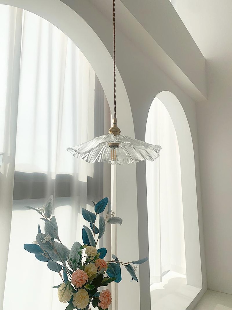 May include: A clear glass pendant light with a ruffled, scalloped shade hangs from a brown cord. The light fixture has a brass-colored base and a visible lightbulb. The background features a white arched window and a vase of flowers.