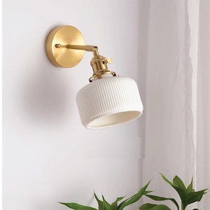 Op de afbeelding: Een witte keramische wandlamp met een gouden metalen basis en arm. De lamp heeft een geribbeld ontwerp en is naar beneden gericht.