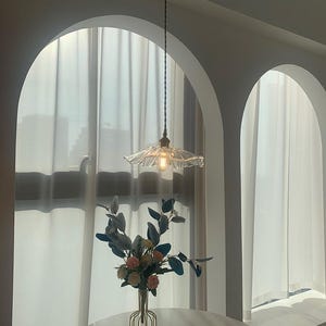 May include: A clear glass pendant light with a ruffled shade hangs from the ceiling. The light fixture is suspended above a round table with a vase of flowers and a film clapperboard. The room has arched doorways and white curtains.