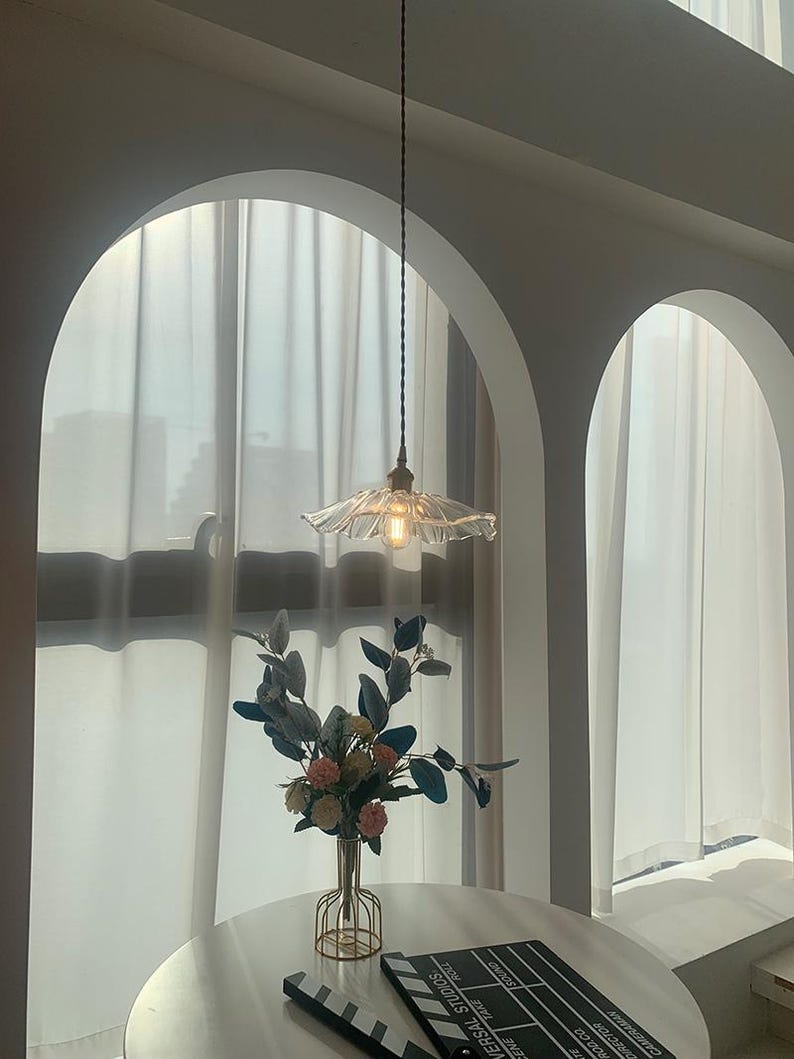 May include: A clear glass pendant light with a ruffled shade hangs from the ceiling. The light fixture is suspended above a round table with a vase of flowers and a film clapperboard. The room has arched doorways and white curtains.