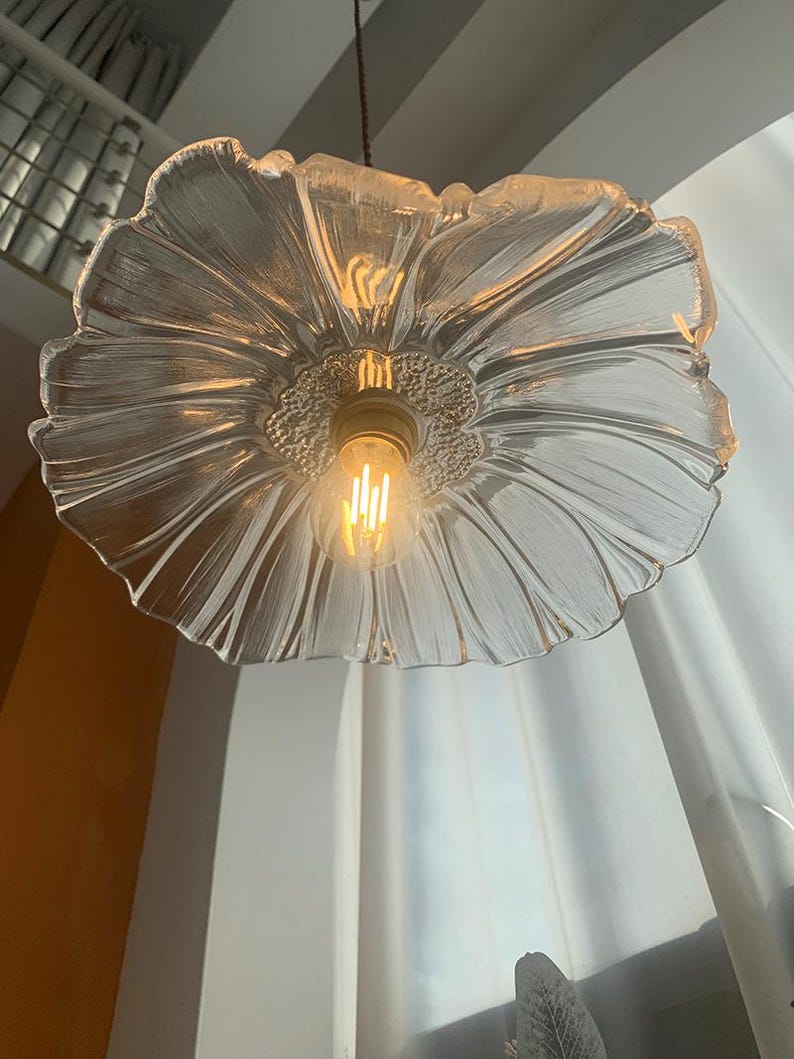 May include: A clear glass pendant light shaped like a flower with a textured, petal-like design. The light fixture has a warm, glowing bulb at its center, suspended from a brown cord. The background includes a white curtain and a glimpse of a window.