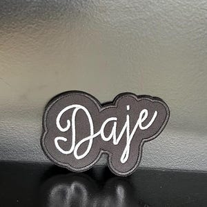 Puede incluir: Un parche negro con la palabra "Daje" en escritura cursiva blanca. El parche tiene forma de nube con un borde negro. Es probable que sea un parche para planchar o coser en ropa o accesorios.