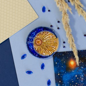 Puede incluir: Un broche de temática celestial con un diseño de luna creciente y sol. El broche es principalmente azul y dorado, con intrincados abalorios y adornos. La luna está adornada con gemas azules, y el sol tiene un centro dorado y radiante. Un accesorio único.