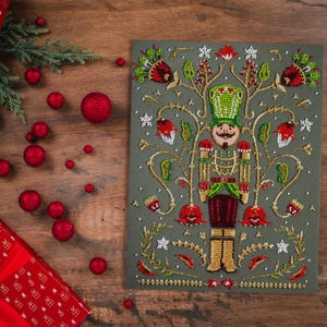 May include: A detailed, beaded Christmas card featuring a nutcracker soldier in green, red, and gold. The card is on a gray background, with red ornaments and a wrapped gift on a wooden surface. Festive holiday decor.