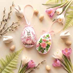 May include: Two decorative Easter egg ornaments with pink ribbon hangers. One egg features a bunny in a pink dress, the other has pink and yellow flowers. White ceramic bunnies, speckled eggs, and purple and white crocus flowers surround the eggs.