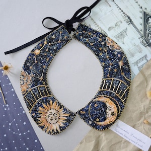 Könnte beinhalten: Eine blaue und goldene Perlenkette mit einem himmlischen Design, das Sonne, Mond und Sterne zeigt. Die Kette ist mit einem schwarzen Band gebunden.