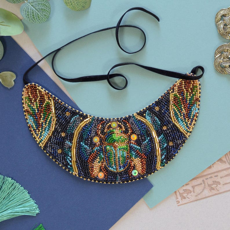 K&ouml;nnte beinhalten: Eine blaue und goldene Perlenkette mit einem Skarab&auml;us-Design. Die Kette hat die Form eines Halbmondes und ist mit einem schwarzen Band versehen.