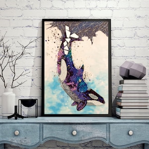May include: Framed artwork featuring a stylized orca whale with a vibrant, beaded design. The whale is depicted in shades of purple, blue, and white against a backdrop of light blue and white. The artwork is framed in black and displayed on a blue surface.
