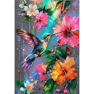 Op de afbeelding: Een diamond painting kunstwerk met een levendige kolibrie in vlucht, omringd door kleurrijke hibiscusbloemen in roze, oranje en witte tinten. Het ontwerp is samengesteld uit kleine, reflecterende kralen op een donkergrijze textuurachtergrond.