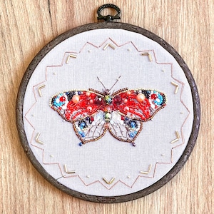 May include: A detailed butterfly embroidery in a wooden hoop. The butterfly is adorned with red, blue, and white beads and sparkling gems. The embroidery is set on a light grey fabric, framed by a pink and gold decorative border.