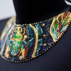 K&ouml;nnte beinhalten: Eine schwarze, blaue, gr&uuml;ne und goldene Perlenkette mit einem gro&szlig;en, detaillierten Design eines Skarab&auml;us.