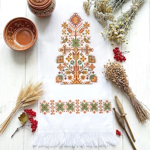 May include: White linen towel with a detailed cross-stitch design in shades of orange, green, and yellow. The towel features a tree-like central motif and a geometric border, with a fringed edge. The image also includes ceramic bowls and wheat.