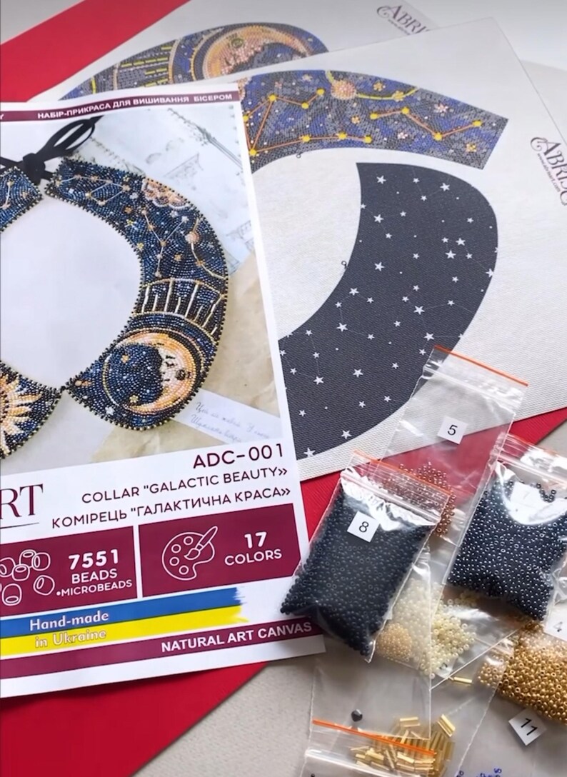 May include: A beading kit for a collar with a celestial theme. The kit includes a canvas, beads in various colors, and instructions. The collar design features a crescent moon, stars, and a sun. The kit is labeled "ADC-001 Collar "Galactic Beauty" Komirets "Galaktichna Krassa" 7551 Beads Microbeads 17 Colors Natural Art Canvas"