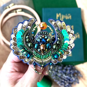 May include: A handcrafted, beaded brooch in the shape of a scarab beetle. The brooch features a blue felt base, adorned with teal sequins, gold beads, and various shades of blue and green beads. An orange crystal sits atop the beetle.