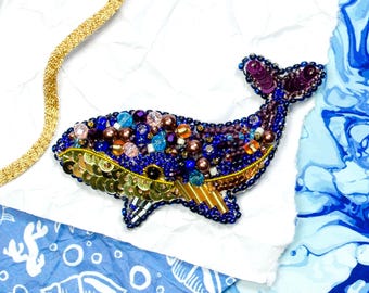 Op de afbeelding: Een handgemaakte walvisbroche, versierd met blauwe, gouden en paarse kralen en pailletten. Het lichaam van de walvis is diepblauw, met een gouden omtrek en een staart van paarse pailletten. De broche ligt op een wit oppervlak met een gouden ketting.