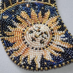 May include: A close-up of a beaded necklace with a crescent moon design. The beads are black, gold, white, and blue. The moon is made of white beads and has a gold outline.