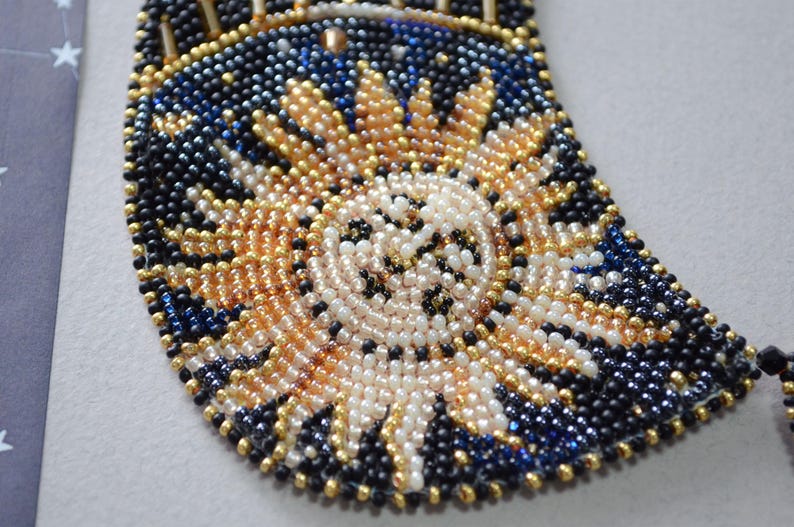 May include: A close-up of a beaded necklace with a crescent moon design. The beads are black, gold, white, and blue. The moon is made of white beads and has a gold outline.