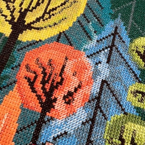 May include: Close-up of a colorful cross-stitch pattern featuring three trees with black trunks and different colored leaves. The trees are stitched on a blue background with a green and yellow background in the upper right corner.