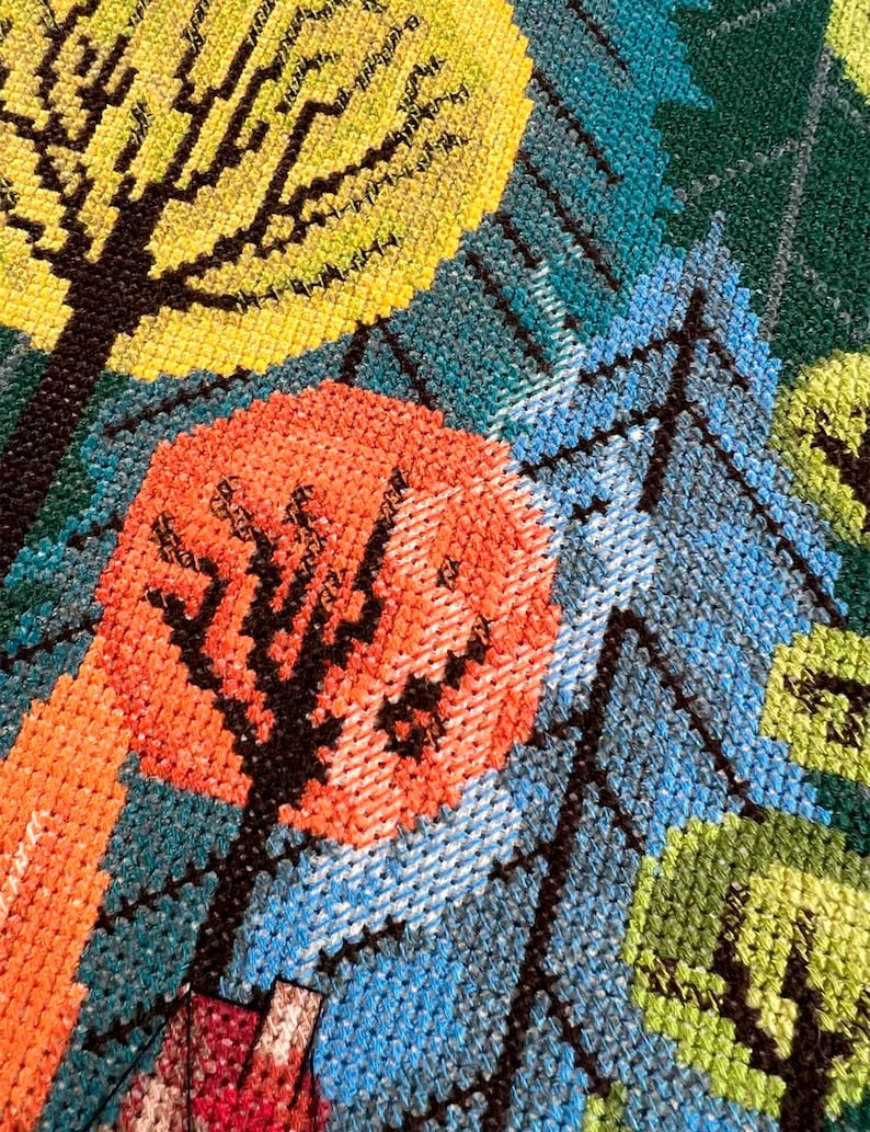 May include: Close-up of a colorful cross-stitch pattern featuring three trees with black trunks and different colored leaves. The trees are stitched on a blue background with a green and yellow background in the upper right corner.