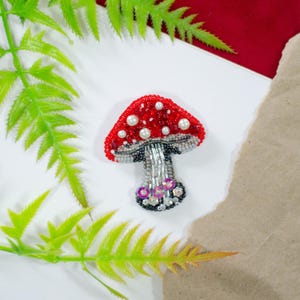 Op de afbeelding: Handgemaakte paddenstoelbroche met een rode, met kralen bezette kap versierd met witte parels. De steel is zilver en grijs, met een zwarte en paarse kralenbasis. De broche staat tegen een witte en rode achtergrond met groen gebladerte.