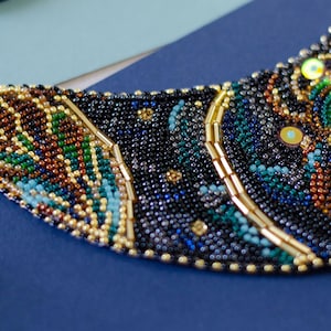 K&ouml;nnte beinhalten: Nahaufnahme einer Perlenkette mit einem geometrischen Muster. Die Kette besteht aus kleinen schwarzen, blauen, gr&uuml;nen und goldenen Perlen.