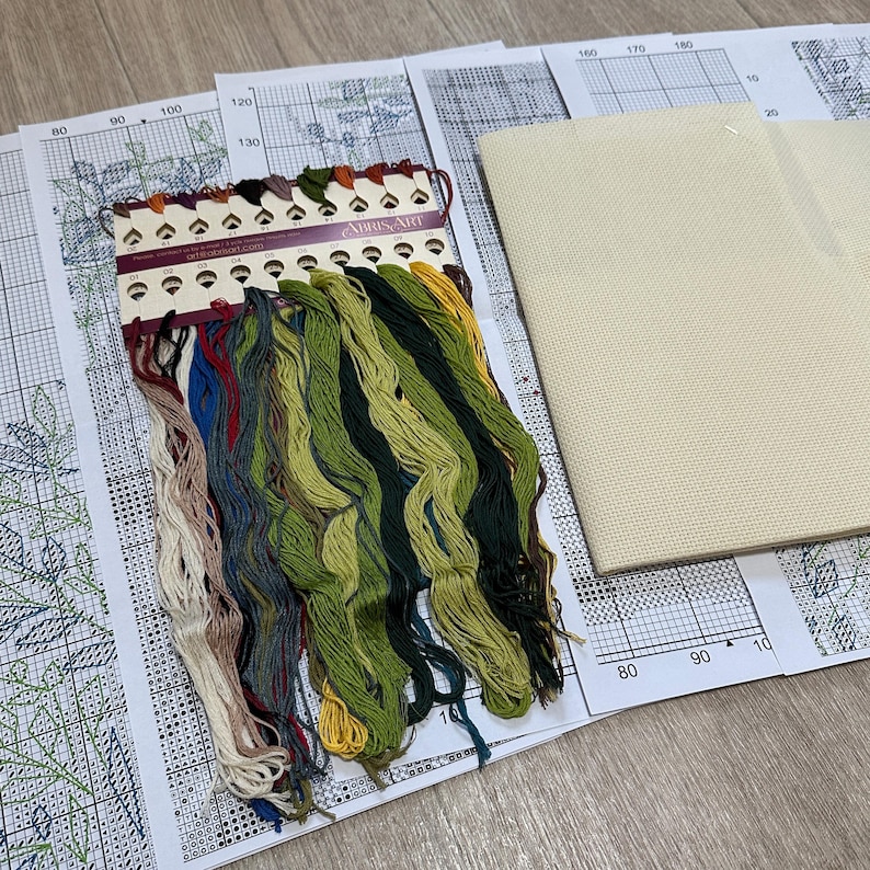May include: A cross stitch kit with a variety of colored threads, a printed pattern, and a piece of Aida cloth. The threads are organized on a card labeled "Abris Art".