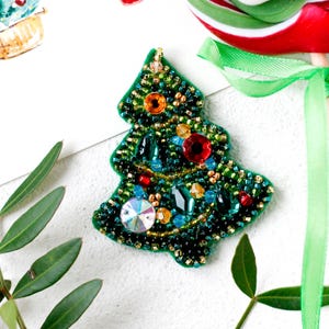 May include: A handcrafted Christmas tree brooch, decorated with green beads and colourful gemstones. The brooch features jewel-toned accents, including red, blue, and orange, and a large, iridescent crystal. The tree is outlined in gold.