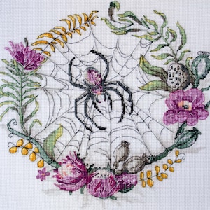 Könnte beinhalten: Eine schwarze Spinne sitzt in der Mitte eines weißen Netzes, umgeben von lila Blumen und grünen Blättern. Die Spinne hat einen rosa Körper und schwarze Beine.