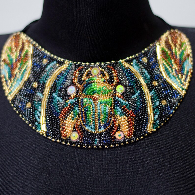 K&ouml;nnte beinhalten: Eine schwarze Perlenkette mit goldener Einfassung und einem gro&szlig;en, bunten Skarab&auml;us-Design in der Mitte.