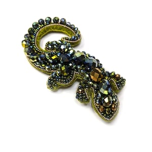 Pode incluir: Broche artesanal em forma de lagarto estilizado. O broche apresenta contas iridescentes verdes, azuis e douradas, fixadas sobre uma base de feltro verde. O design inclui uma cauda enrolada e detalhes de contas.