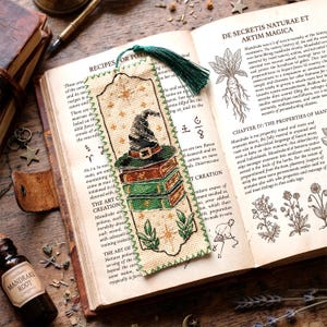Puede incluir: Un marcapáginas rectangular de punto de cruz sobre tela beige con borde verde y una borla verde oscuro. El diseño muestra una pila de libros verdes coronada por un sombrero de bruja, rodeada de pequeñas estrellas doradas y hojas verdes.