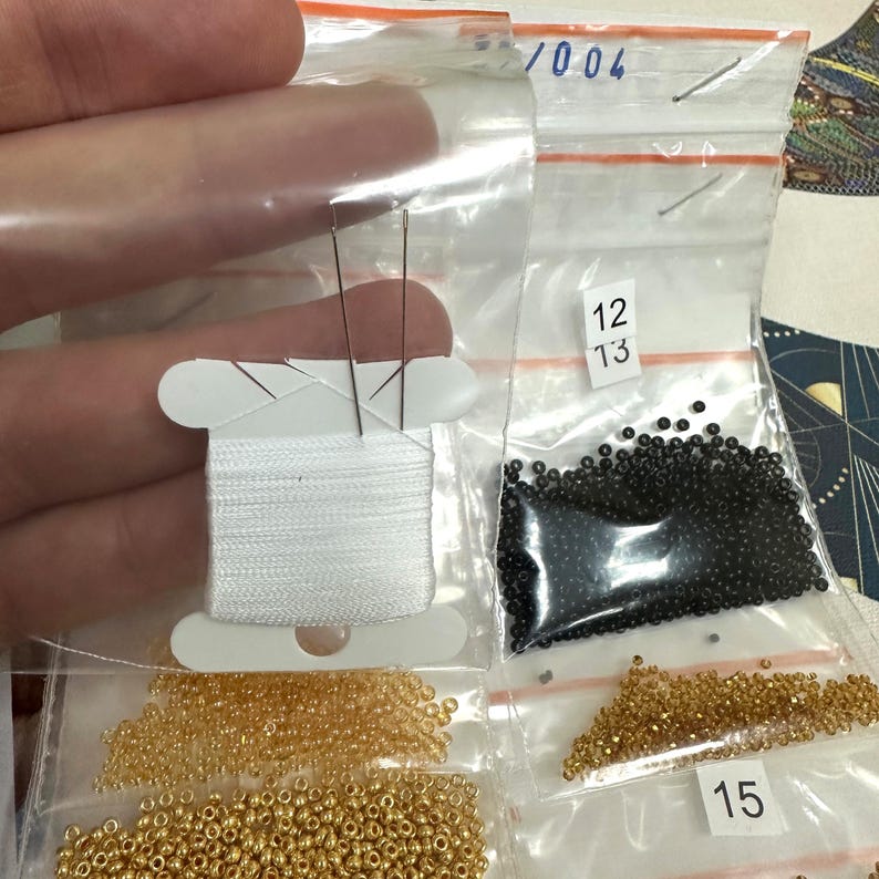 K&ouml;nnte beinhalten: Nahaufnahme eines Perlen-Sets mit wei&szlig;em Garn, schwarzen Perlen und goldenen Perlen in separaten Plastikt&uuml;ten. Die Beutel sind mit den Nummern 12, 13 und 15 beschriftet.