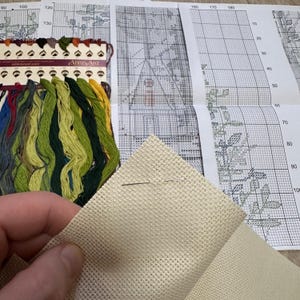 May include: A close-up of a beige Aida cloth with two needles on top. The cloth is used for cross-stitch embroidery. The cloth is next to a printed cross-stitch pattern with a grid and numbers.
