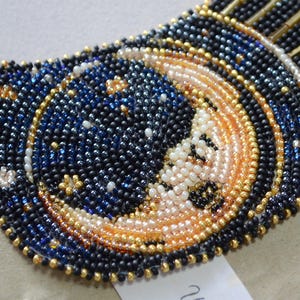 May include: A close-up of a beaded necklace with a crescent moon design. The moon is made of white and gold beads, and the background is a dark blue with gold stars.