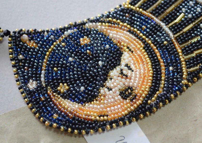 May include: A close-up of a beaded necklace with a crescent moon design. The moon is made of white and gold beads, and the background is a dark blue with gold stars.