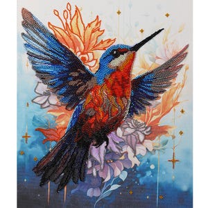 Op de afbeelding: Een levendige, met kralen bewerkte kolibrie met uitgespreide vleugels, met een spectrum van blauw, rood en oranje. Het kunstwerk bevat bloemaccenten in oranje, wit en paars, tegen een blauwe achtergrond.