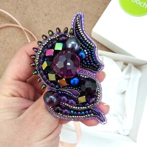 Op de afbeelding: Handgemaakte broche met kralen en kristallen in paarse, zwarte en iriserende kleuren. De broche heeft een complex ontwerp met kralen en kristallen in verschillende maten en vormen, wat een sprankelend accessoire oplevert.
