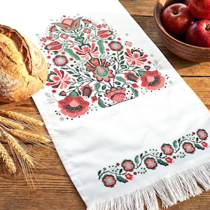 May include: White embroidered table runner with a floral design in red, green, and pink. The runner features a fringed edge and is displayed on a wooden table. A loaf of bread and a bowl of apples are also visible.