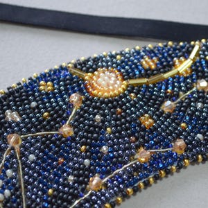 May include: A close-up of a beaded necklace with a constellation design. The beads are black, blue, and gold, and the constellation is made of silver wire. The necklace is a beautiful and unique piece of jewelry.