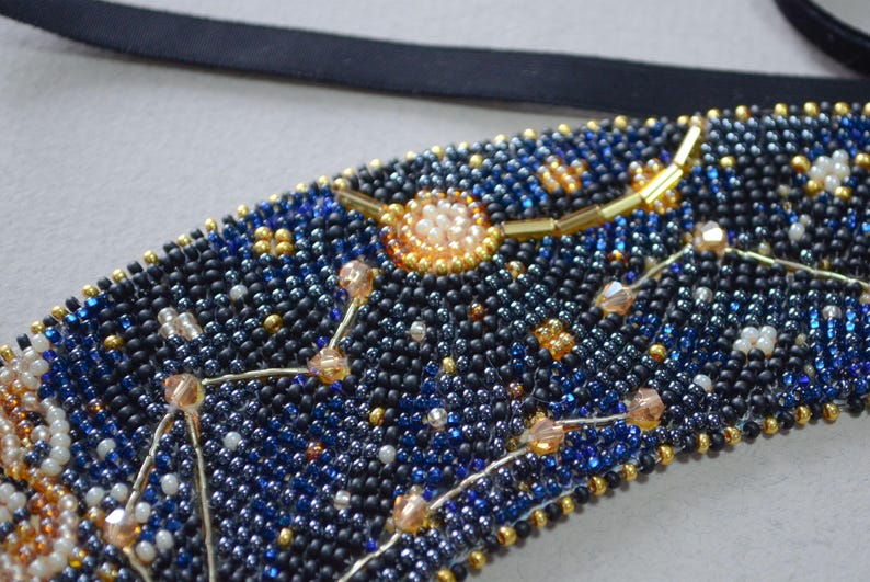 May include: A close-up of a beaded necklace with a constellation design. The beads are black, blue, and gold, and the constellation is made of silver wire. The necklace is a beautiful and unique piece of jewelry.