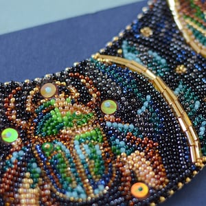K&ouml;nnte beinhalten: Eine Perlenkette in Halbmondform mit verschiedenen Farben wie Schwarz, Braun, Gr&uuml;n und Gold. Die Kette ist mit schillernden Pailletten verziert.