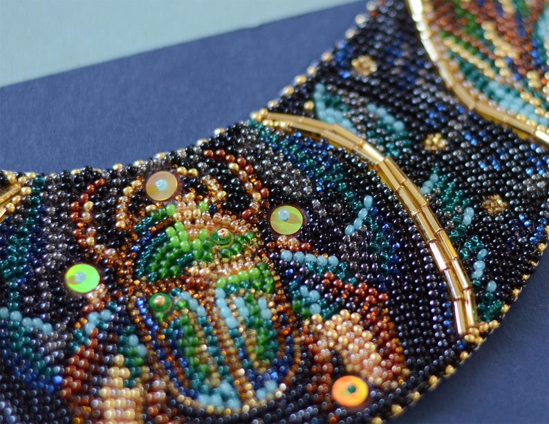 K&ouml;nnte beinhalten: Eine Perlenkette in Halbmondform mit verschiedenen Farben wie Schwarz, Braun, Gr&uuml;n und Gold. Die Kette ist mit schillernden Pailletten verziert.