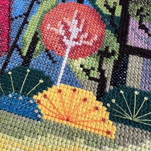 May include: A close-up of a cross-stitch embroidery depicting a stylized forest scene with a bright orange tree, yellow flowers, and green foliage.