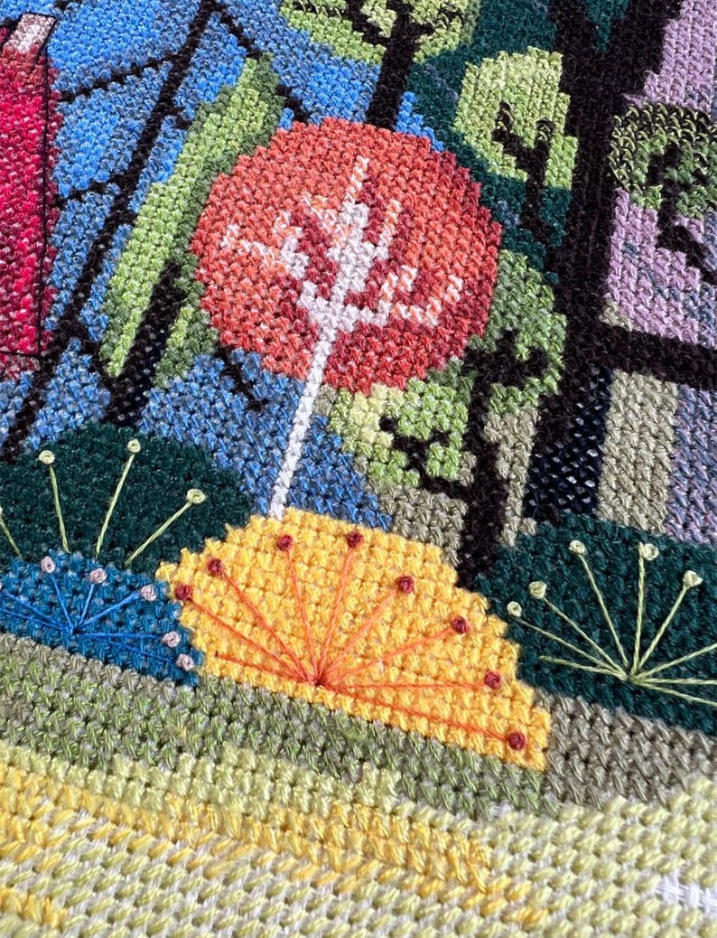 May include: A close-up of a cross-stitch embroidery depicting a stylized forest scene with a bright orange tree, yellow flowers, and green foliage.